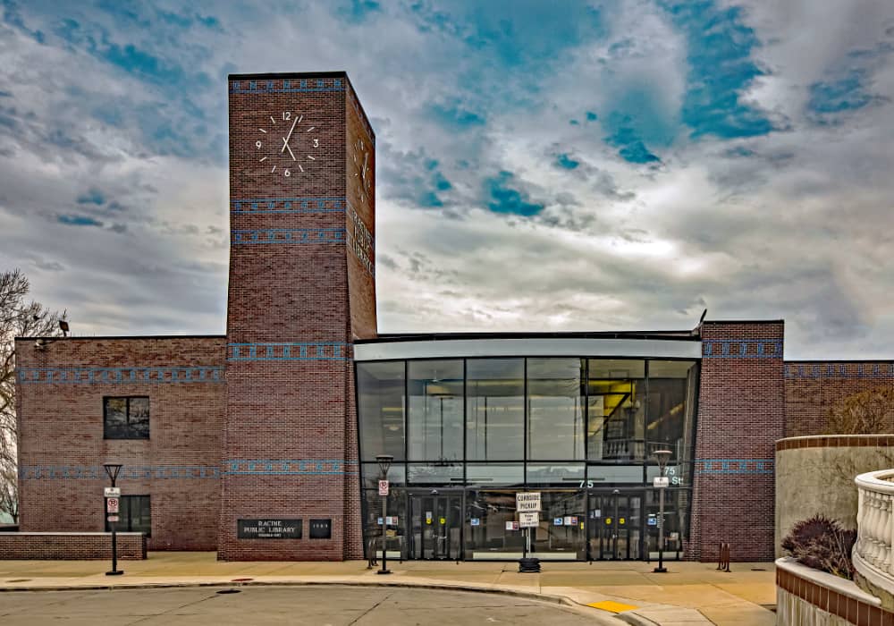 A photo of the library's Library Drive entrance. It's a dark red brick building with aqua blue tile accents and a tall clock tower on the left side of the tall glass windows and doors encasing the front lobby.