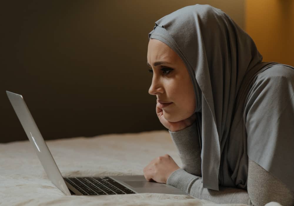 A tan person in a gray headscarf lays on their stomach and props a head in their hand while staring at a laptop screen.