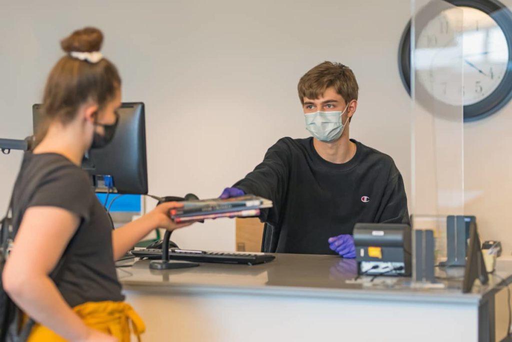 A photo of Matt Klug, a lean, white circulation worker with short brown hair, wearing a mask and blue gloves as he hands a stack of books across a counter to a masked patron.