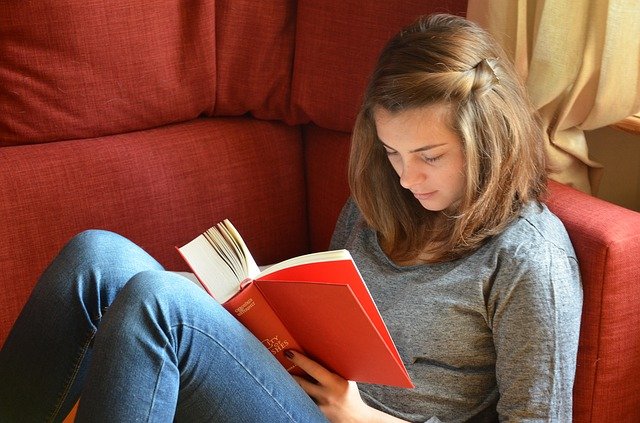 A white person with shoulder-length brown hair sits sideways in an armchair and reads a book.
