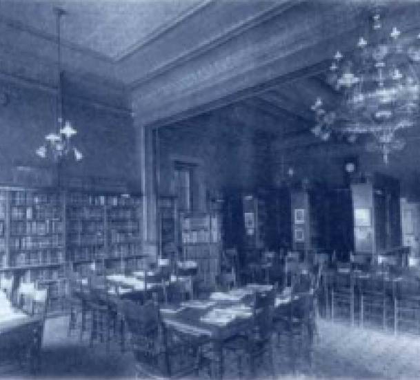 A black and white image of the interior of the Racine Public Library's first location at 245 Main Street in 1897. You can see ornate chandeliers, tall bookshelves and tables with several chairs each.
