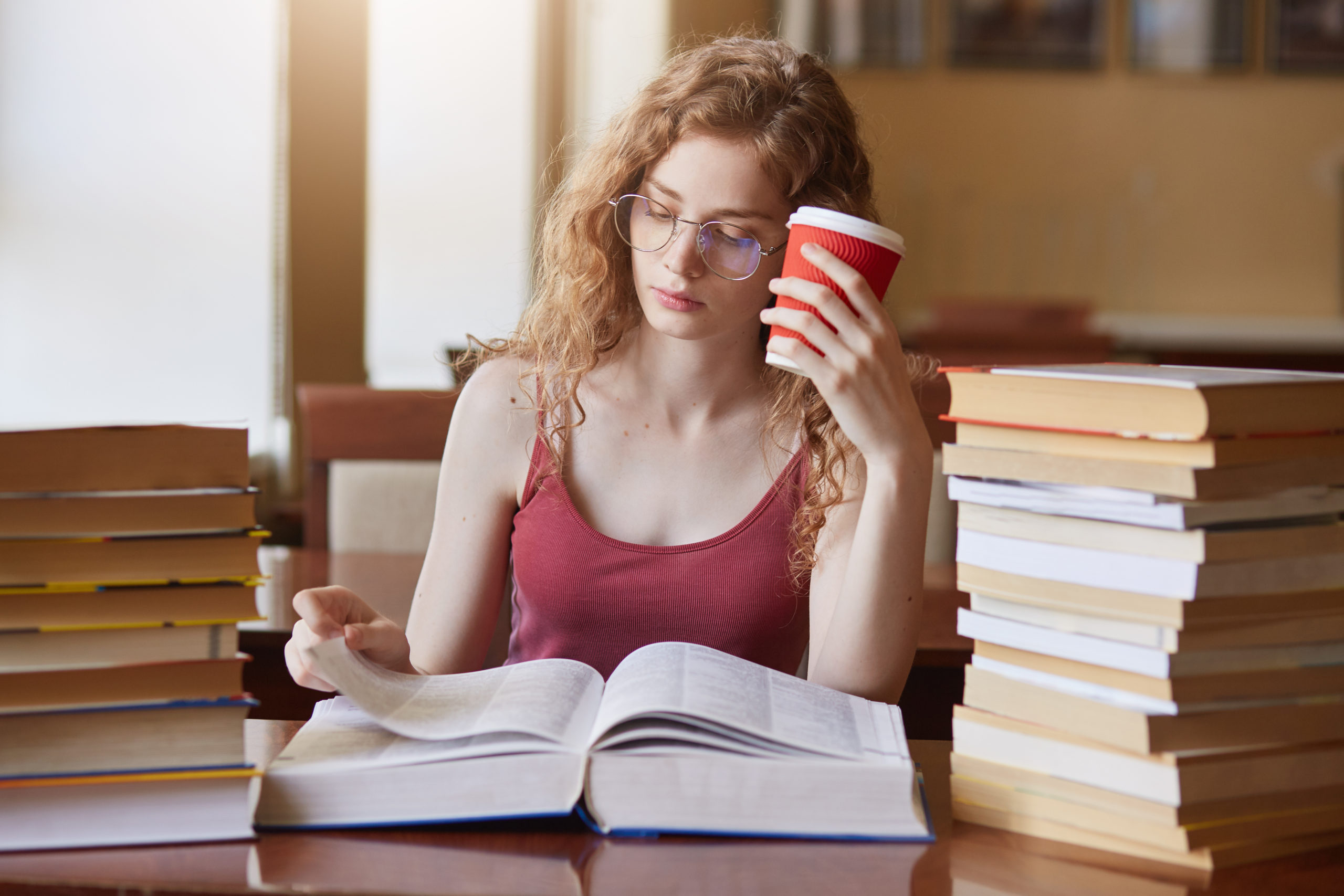 A thin, white redhead with long, curly hair pores over a thick book. They sit between two stacks of more books and hold a coffee cup in their free hand.