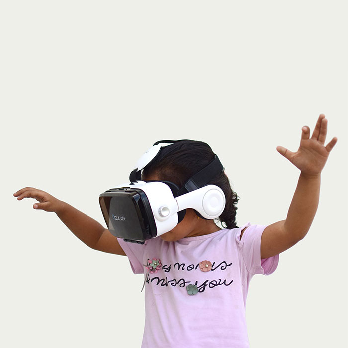 A Brown child with long, curly hair in a ponytail waves their hands in the air and wears a virtual reality mask.