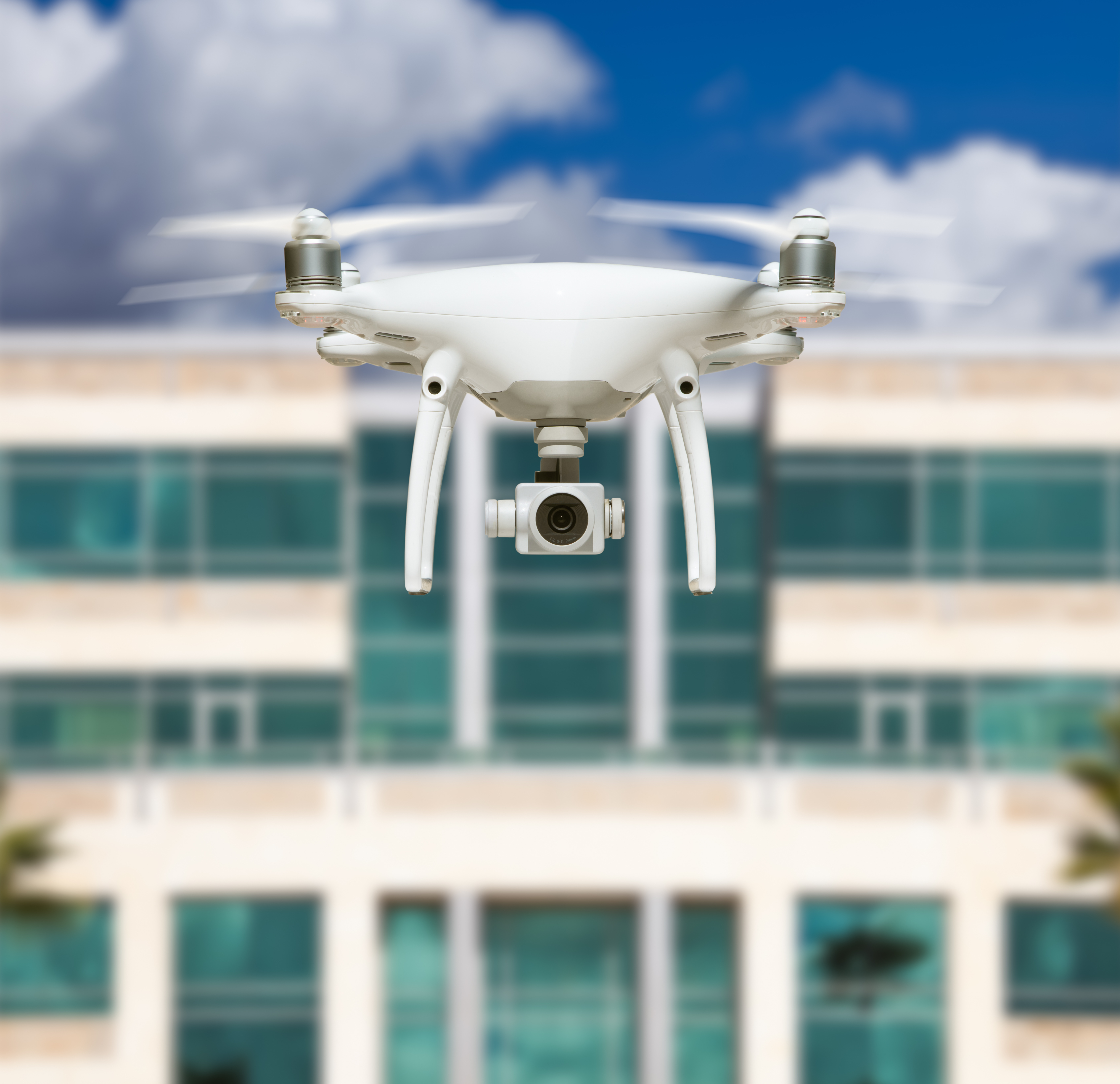 A white, remote-controlled drone robot hovers in front of a tall building.