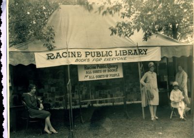 A yellowed, black and white photo of a tent with a sign that says "Racine Public Library. books for everyone." Under the tent are three adults and a child running the booth. A sign underneath says "Racine Public Library. All kinds of books for all kinds of people." On the back wall of the tent are several sheets of paper, perhaps pages from different books, with different photos and text on them.