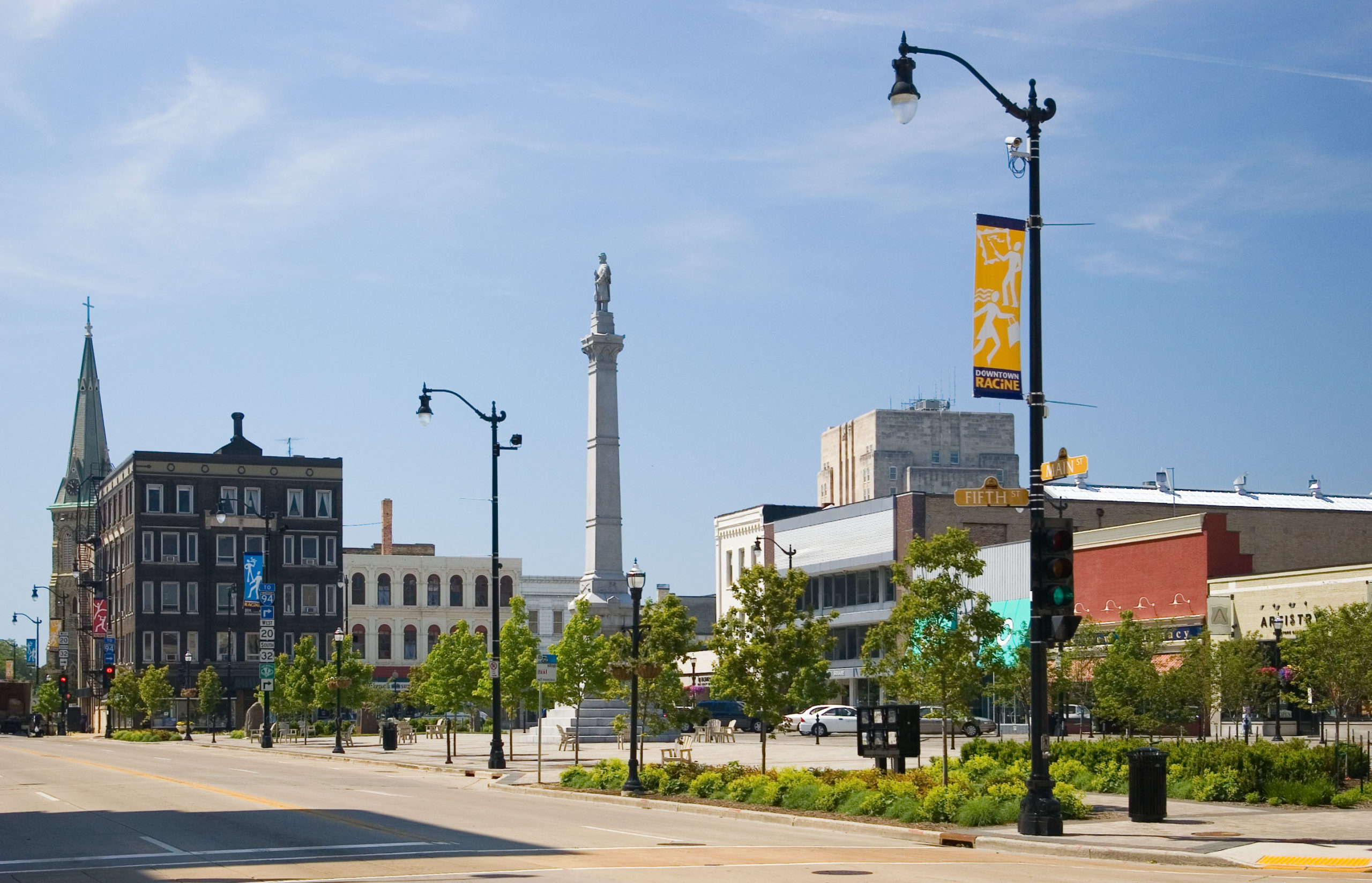 A photo of downtown Racine's main street square. You can see the monument in the middle, framed by sidewalk, benches, streetlamps, trees and local businesses.