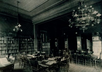 A black and white photo of the interior of the first Racine Public Library building. The room has several tall bookshelves along the back and is full of dining tables surrounded by chairs, with books covering each table. Tall, ornate chandeliers hang from the high ceilings, and the ceilings have several inlets with every surface being covered in ornate details.