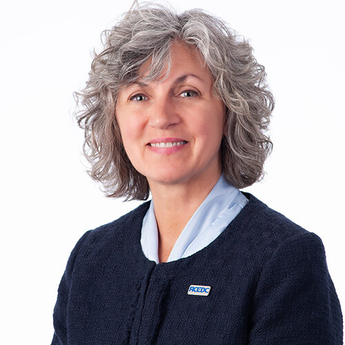 A photo of Jenny Trick, a thin, white woman. She's wearing a navy blue RCEDC pullover, and her short, gray hair is styled into curls around her head.