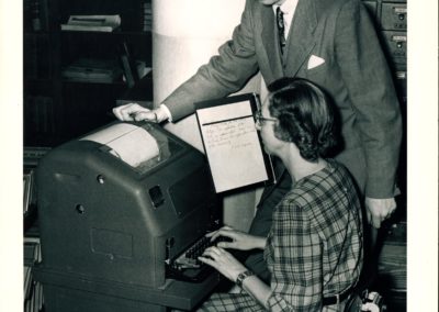 A black and white photo of a person sitting at what looks like an extremely bulky typewriter, with the keyboard attached to a larger machine the size of a UPS mailbox. A sheet of paper is mounted on a stand off of the machine for reference. While the person types on the teletype, another person leans over the machine to observe.