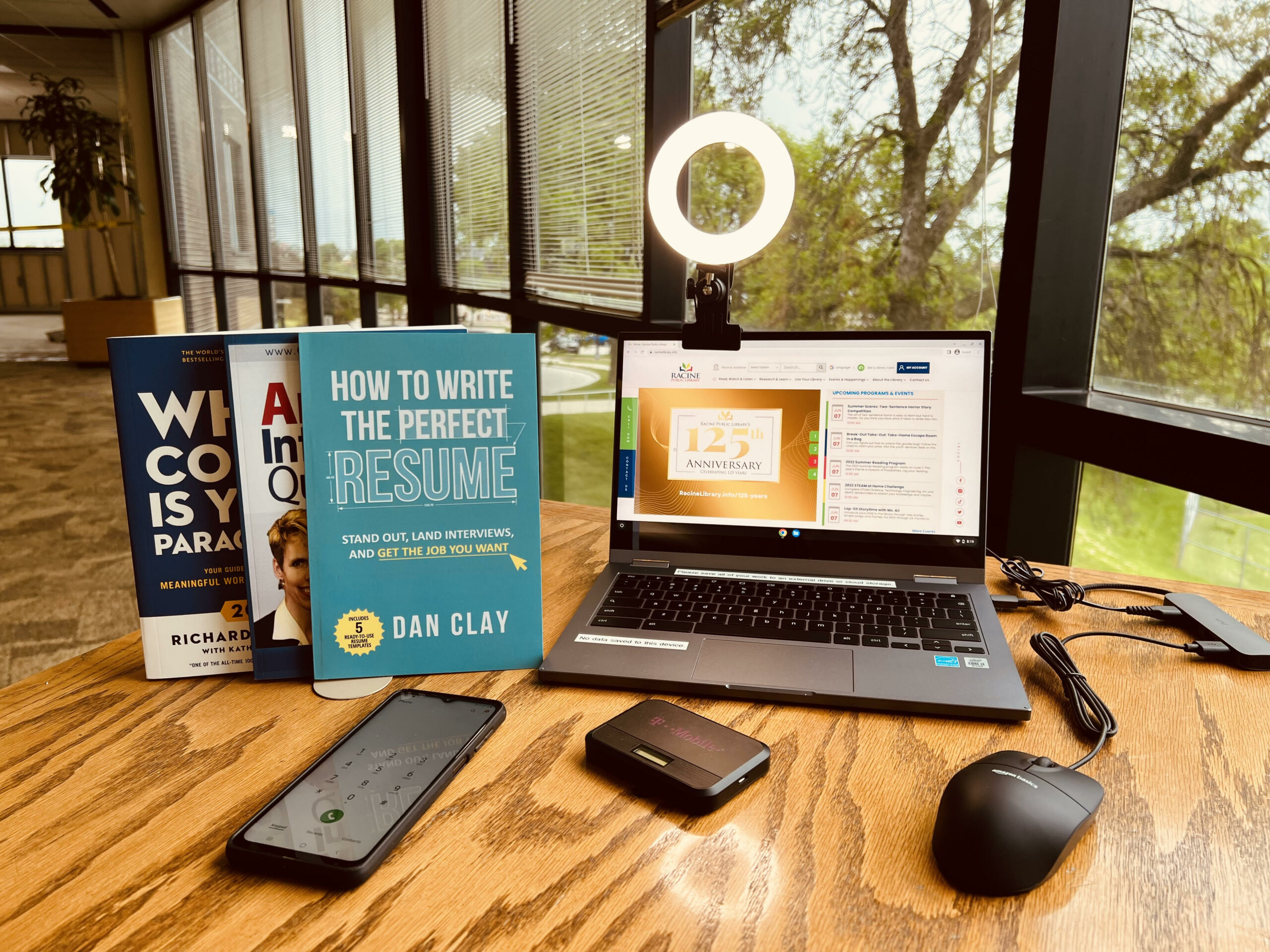 A photo of the items available in the library's career connection kit. There's a laptop with a ring light clipped onto it, a USB port for the computer connecting it to the ring light and a wired mouse, an Android smartphone, a T-Mobile wireless hotspot, and three job-hunting books, including How to Write the Perfect Resume by Dan Clay.