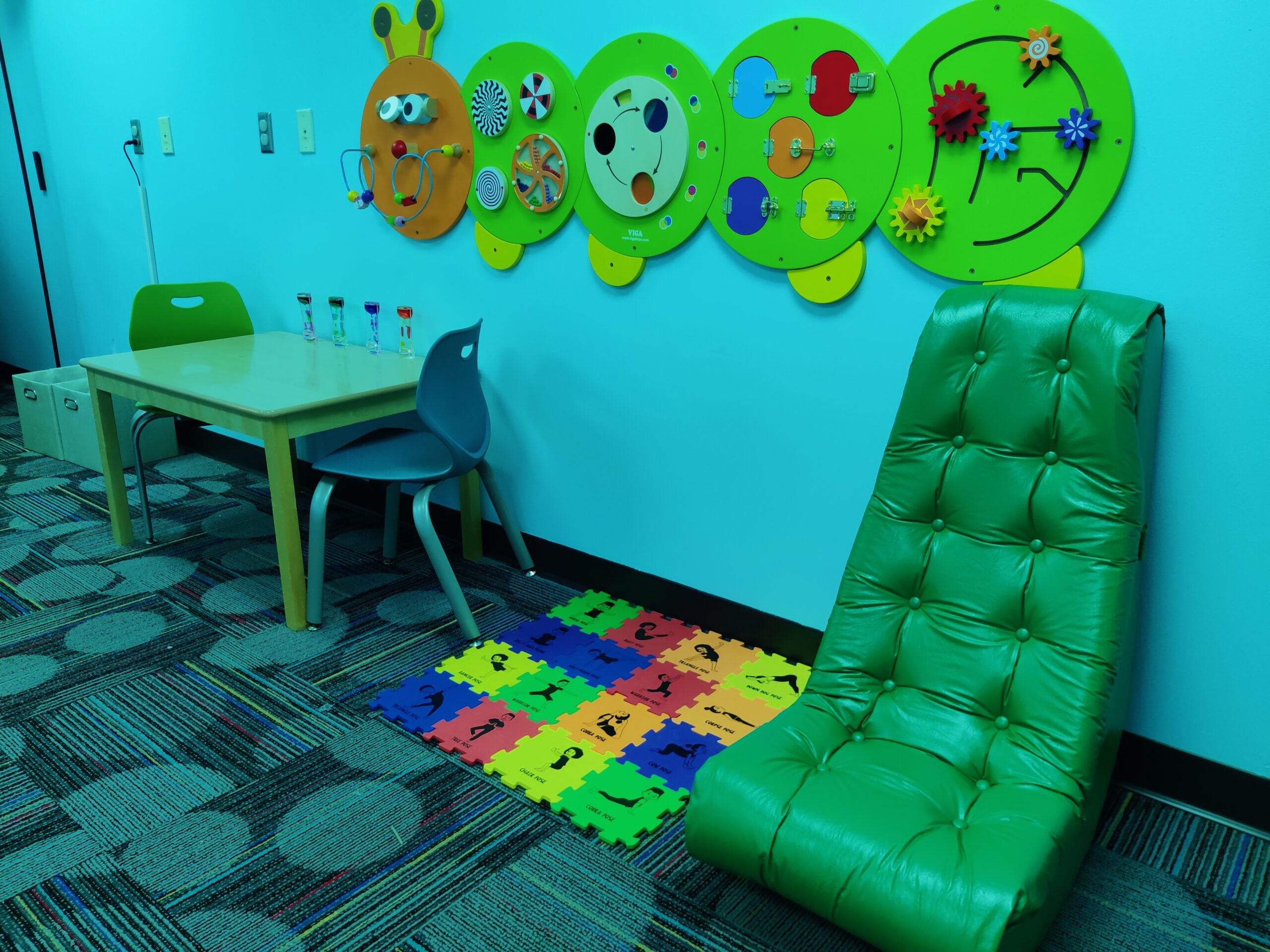 A photo of the sensory room in dim, green-toned lighting. This angle of the room shows a wall-mounted caterpillar sensory game, a table with liquid hourglasses on it, a floor rocking chair, and a foam puzzle mat with different exercise prompts on it.