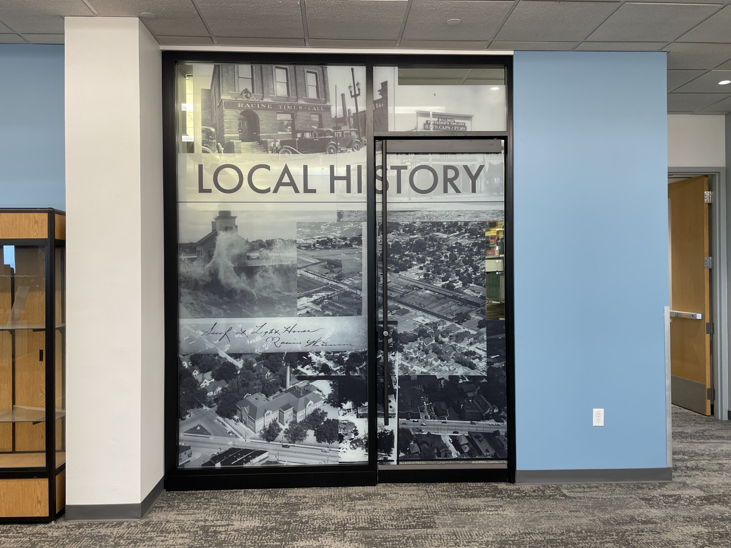 A photo of the doorway to the library's renovated History room. The doorway looks like a large panorama of black and white photography featuring historic buildings and old photography from Racine, with a label across the top that says "Local History." A section of the photo wall is the door, which opens into the history room.