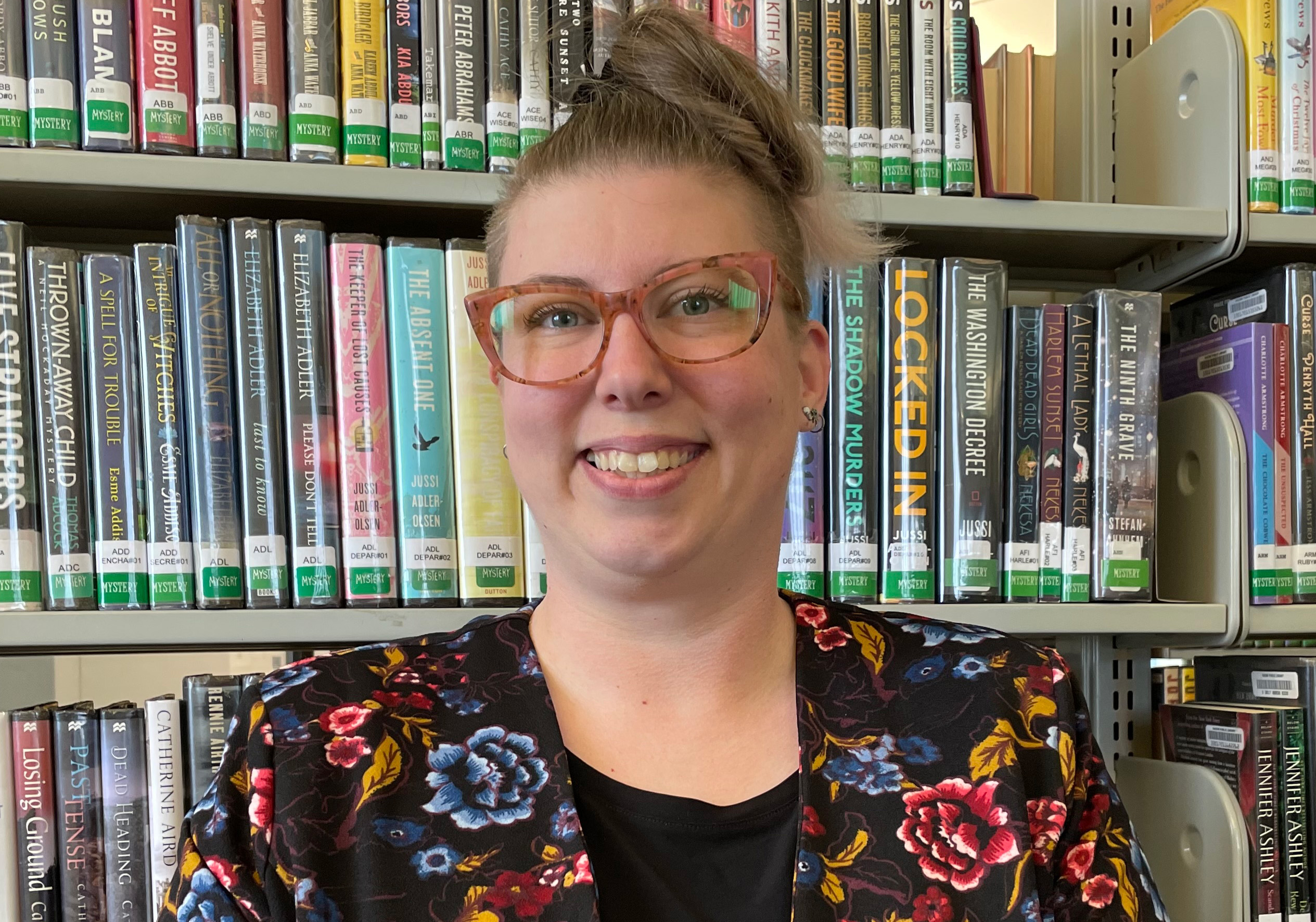 25-05-19 Becky Crowley Portrait - rectangular, cropped Becky Crowley is a straight-sized white person with their hair in a loose bun on top of their head, pink cat-eye glasses and a black floral jacket.